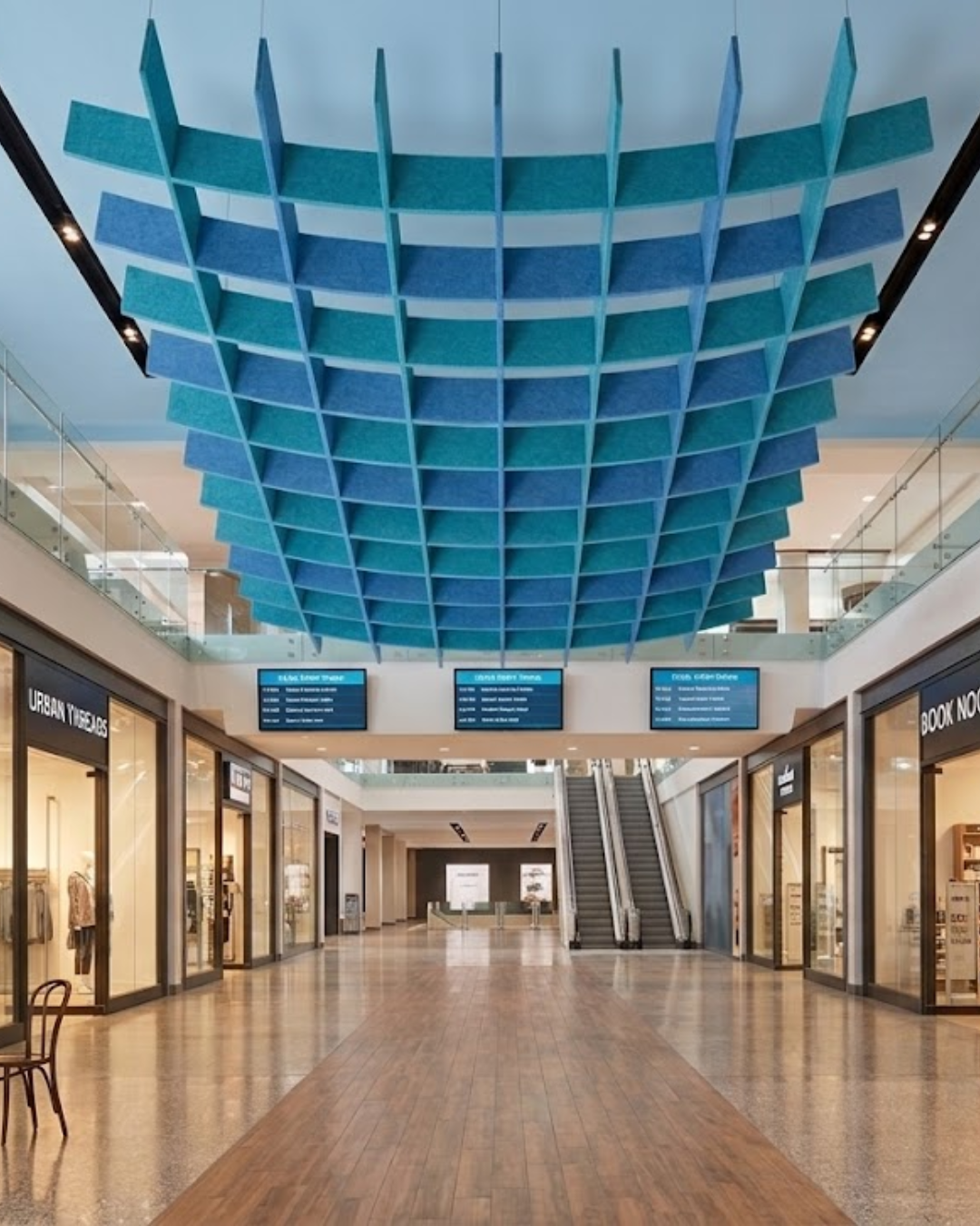 A large suspended Acoustic Baffle ceiling panels system in shades of blue and teal installed in a modern shopping mall corridor with storefronts, escalators, and a polished floor.