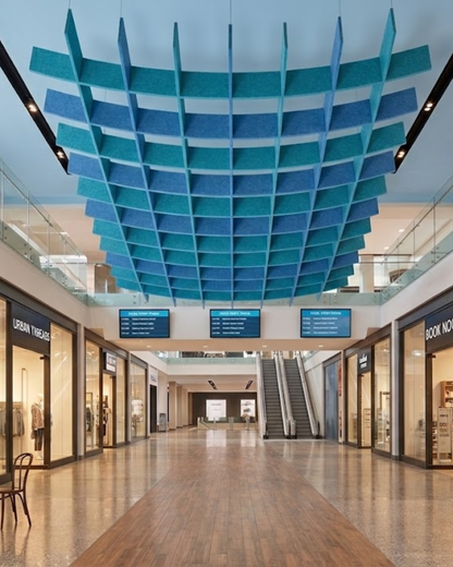 A large suspended Acoustic Baffle ceiling panels system in shades of blue and teal installed in a modern shopping mall corridor with storefronts, escalators, and a polished floor.
