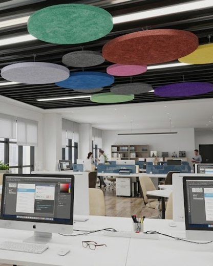A modern office interior with several large, round, suspended Acoustic Baffle ceiling panels in various colors including green, red, blue, pink, yellow, and purple. In the foreground, white desks feature computer monitors, keyboards, and eyeglasses.