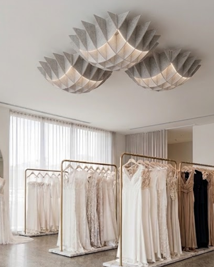 A modern bridal boutique interior with three sculptural paper lantern Acoustic Baffle ceiling panels lights glowing above racks of white, cream, and beige wedding dresses.