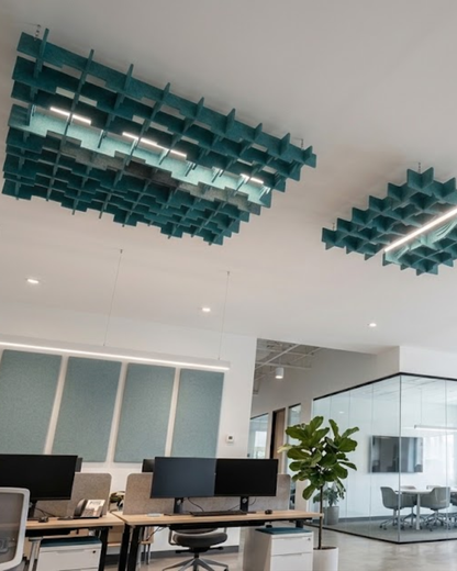 Modern office interior with two large teal geometric Acoustic Baffle ceiling panels and matching wall panels, office desks with computer monitors, a potted plant, and glass meeting rooms.