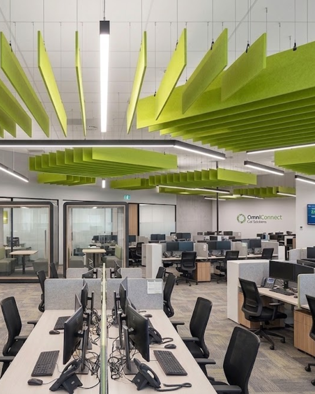A modern open-plan office interior with rows of desks, computer monitors, office chairs, and bright lime green Acoustic Baffle ceiling panels suspended from the ceiling.